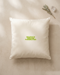 A square cream organic cotton cushion lies on a smooth beige plaster surface, touched by soft shadows of sunlight. In the center, bold lime green lettering reads “Believe in endless possibilities.” In the top right corner, a sprig of dried white sage rests beside a small clear quartz crystal.