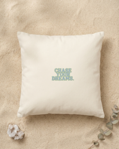 A square cream organic cotton cushion rests on fine golden sand. In the center, bold dusty blue lettering with a thin green outline reads “Chase your dreams.” A small white quartz crystal sits in the bottom left corner, and a sprig of silver green eucalyptus reaches in from the bottom right.