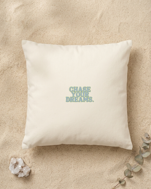 A square cream organic cotton cushion rests on fine golden sand. In the center, bold dusty blue lettering with a thin green outline reads “Chase your dreams.” A small white quartz crystal sits in the bottom left corner, and a sprig of silver green eucalyptus reaches in from the bottom right.