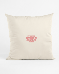 A square cream organic cotton cushion stands upright against a plain white studio background. In the center, bold soft coral pink lettering with a darker pink outline reads “It gets better, it always does.”