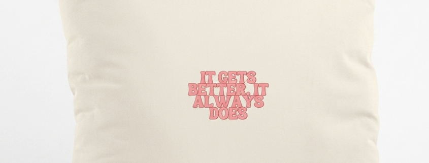 A square cream organic cotton cushion stands upright against a plain white studio background. In the center, bold soft coral pink lettering with a darker pink outline reads “It gets better, it always does.”