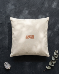 A square cream organic cotton cushion lies on a textured dark slate surface, dappled with soft light and leaf shadows. In the center, bold terracotta lettering reads “Make it happen.” A small clear quartz crystal sits in the bottom left corner, and a sprig of silver green eucalyptus reaches in from the bottom right.