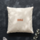 A square cream organic cotton cushion lies on a textured dark slate surface, dappled with soft light and leaf shadows. In the center, bold terracotta lettering reads “Make it happen.” A small clear quartz crystal sits in the bottom left corner, and a sprig of silver green eucalyptus reaches in from the bottom right.