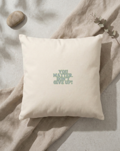 A square cream organic cotton cushion rests on a draped taupe linen cloth over a pale grey stone surface, with soft leaf shadows falling across the scene. In the center, bold dusty blue lettering with a thin green outline reads “You matter. Don’t give up!” A smooth river pebble sits in the top left corner, and a sprig of eucalyptus reaches in from the top right.