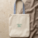 A cream organic cotton tote bag lies flat on a beige travertine surface, partly resting on a crumpled taupe linen cloth. Its handles are woven in a blue and white diamond pattern. In the center, small bold lettering in soft mint green with a blue outline reads “You’ve got this!”